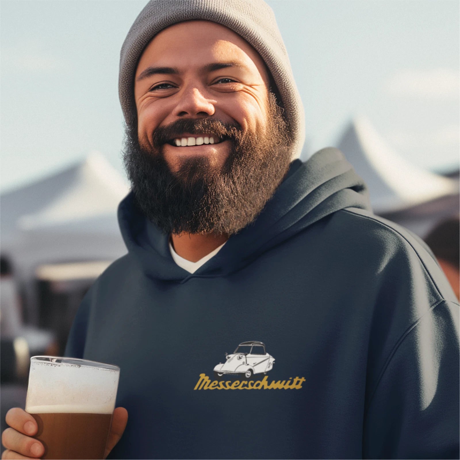 Man wearing a hoodie with a logo, holding a beer, with tents in the background