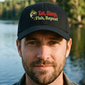 A black baseball cap featuring a front graphic with a green fish illustration and the text 'Eat, Sleep, Fish, Repeat' in red and yellow-green lettering, shown on a plain white background.