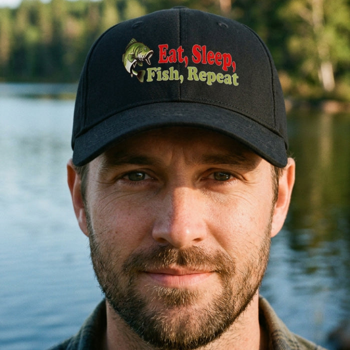A black baseball cap featuring a front graphic with a green fish illustration and the text 'Eat, Sleep, Fish, Repeat' in red and yellow-green lettering, shown on a plain white background.