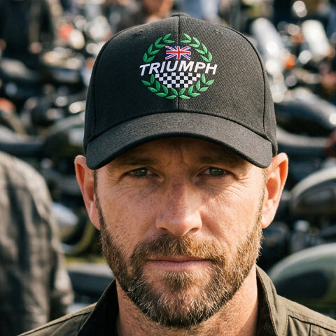 A black baseball cap with a custom printed Triumph logo on the front, featuring a green laurel wreath, a Union Jack flag, and a checkered pattern. The brim has a placeholder text 'Custom Text Here' in white script.