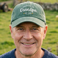 A green-washed baseball cap with white printed text reading 'PROMOTED TO Grandpa EST. 2026' on the front and 'Custom Text' on the brim, shown on a plain white background.