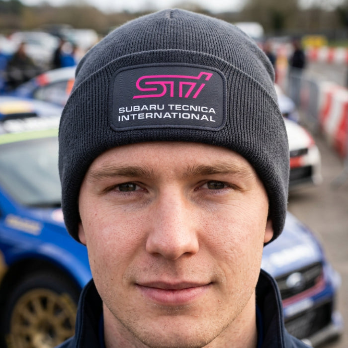 A close-up portrait of a man wearing a dark gray ribbed beanie with a rectangular patch that reads 'SUBARU TECNICA INTERNATIONAL' in white text with a pink stylized logo. A blue Subaru rally car is blurred in the background.