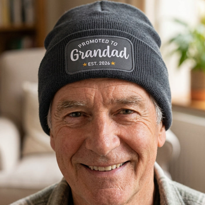 A dark gray ribbed knit beanie with a folded cuff, featuring a rectangular black patch on the front with the white text 'PROMOTED TO Grandad' and 'EST. 2026' with small gold stars.