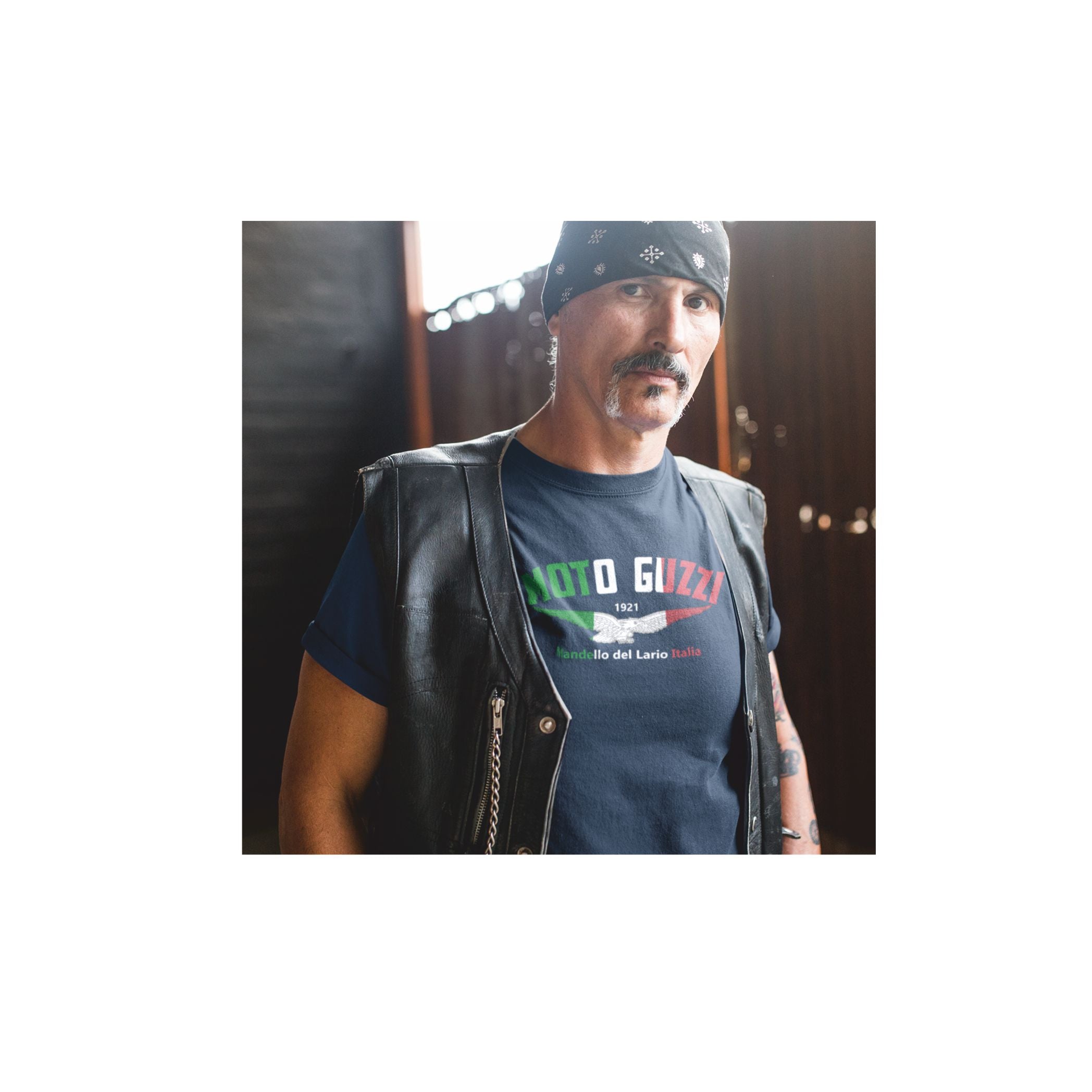 Man wearing a black leather vest over a dark t-shirt with a logo, standing against a blurred background.