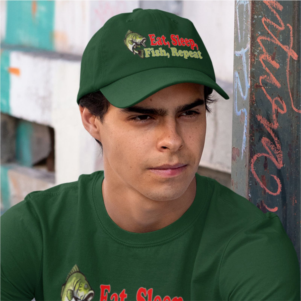A man wearing a dark green baseball cap with an embroidered graphic of a fish and the text 'Eat, Sleep, Fish, Repeat' in red and yellow-green lettering. He is also wearing a matching dark green t-shirt, set against a blurred urban background.