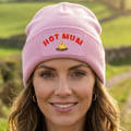 A woman wearing a light pink knit beanie with a patch that reads 'HOT MUM' in red letters above a small campfire graphic. The background is a softly blurred green outdoor landscape.