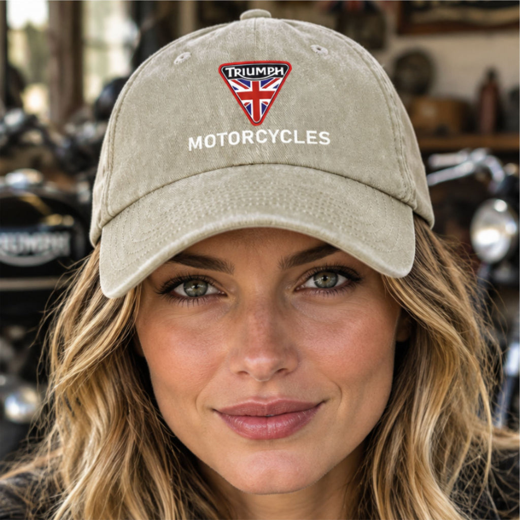 A woman wearing a beige baseball cap with a red, white, and blue embroidered Triumph Motorcycles shield logo on the front, set against a blurred motorcycle shop background.