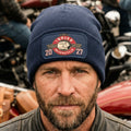 A navy blue ribbed knit beanie with a folded cuff, featuring a rectangular printed  patch on the front. The patch displays a circular emblem with the words 'Indian Riders Group Motorcycle 2027' and a Native American profile illustration.