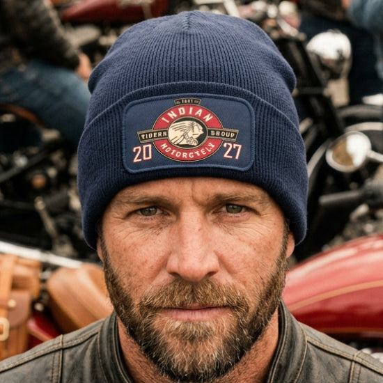 A navy blue ribbed knit beanie with a folded cuff, featuring a rectangular printed  patch on the front. The patch displays a circular emblem with the words 'Indian Riders Group Motorcycle 2027' and a Native American profile illustration.