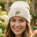 A cream-colored ribbed knit beanie hat featuring a rectangular printed patch on the front cuff. The patch displays the text 'Ask The Expert' in orange script above an illustration of a wooden wheelbarrow filled with purple, yellow, and green flowers.