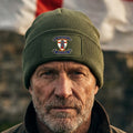 An olive green ribbed knit beanie hat with a folded cuff, featuring a rectangular patch on the front with a British Royal Marines crest and the motto 'COME THE WORLD AGAINST THEM' and 'ENGLAND WILL SHALL STAND'.