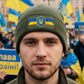 An olive green ribbed knit beanie with a folded cuff, featuring a rectangular patch on the front with a blue and yellow Ukrainian flag design and a central gold trident emblem.