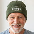 An olive green ribbed knit beanie hat with a folded cuff, featuring a rectangular embroidered patch that reads 'PROMOTED TO Grandpa EST. 2026' in white and yellow.