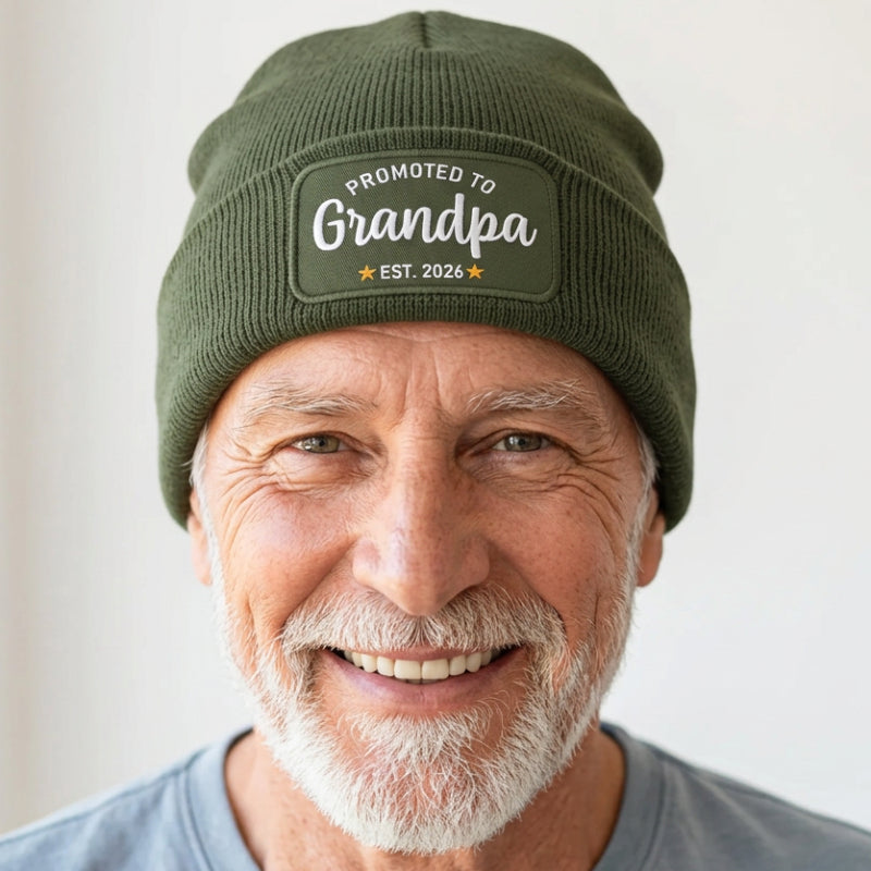 An olive green ribbed knit beanie hat with a folded cuff, featuring a rectangular embroidered patch that reads 'PROMOTED TO Grandpa EST. 2026' in white and yellow.