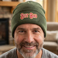 An olive green ribbed knit beanie hat featuring a rectangular printed patch on the front cuff with the words 'BEST DAD' in red with a gold outline.