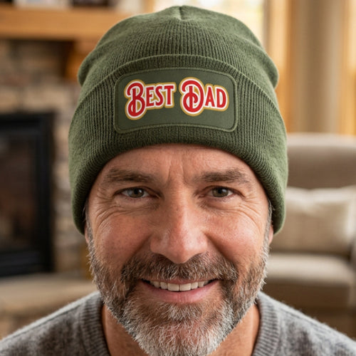 An olive green ribbed knit beanie hat featuring a rectangular printed patch on the front cuff with the words 'BEST DAD' in red with a gold outline.