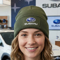 An olive green ribbed knit beanie hat featuring a rectangular patch on the front cuff with the blue and silver Subaru logo and white 'SUBARU' text.