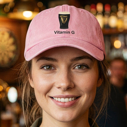 A light pink cotton twill baseball cap featuring a graphic of a black pint glass with a gold harp and the text 'Vitamin G' printed in black below it.