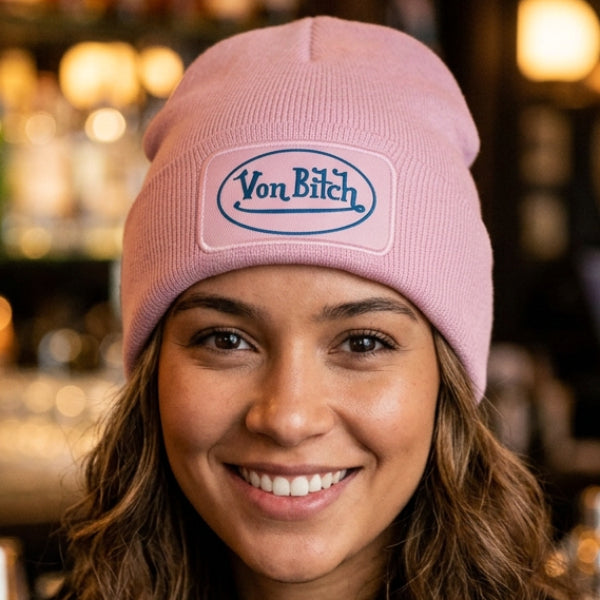A light pink ribbed knit beanie hat featuring a rectangular patch on the front cuff with the text 'Von Bitch' in blue script inside a blue oval.