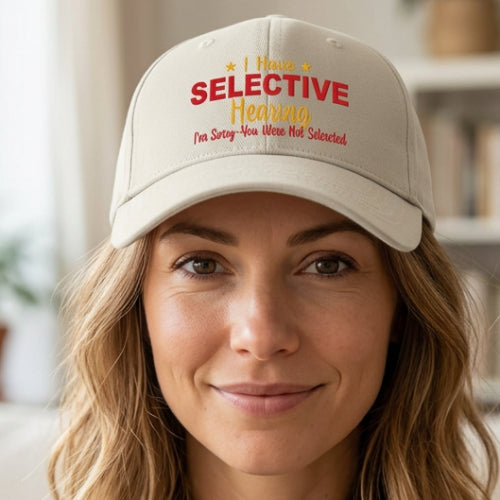 An off-white baseball cap with printed text on the front reading 'I Have SELECTIVE Hearing I'm Sorry You Were Not Selected' in red and yellow lettering.