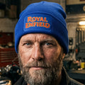 A royal blue knit beanie hat against a light motorcycle background. The front cuff features a rectangular patch with the words 'ROYAL ENFIELD' in orange lettering with a yellow outline.