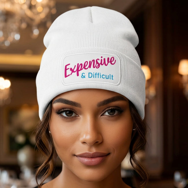 A white ribbed knit beanie hat featuring a white rectangular patch on the front cuff with the text 'Expensive & Difficult' in pink and blue lettering, set against a plain white background.