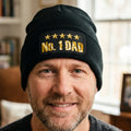 A black ribbed knit beanie hat featuring a rectangular black patch on the front cuff with five gold stars and the text 'No. 1 DAD' in metallic gold lettering.