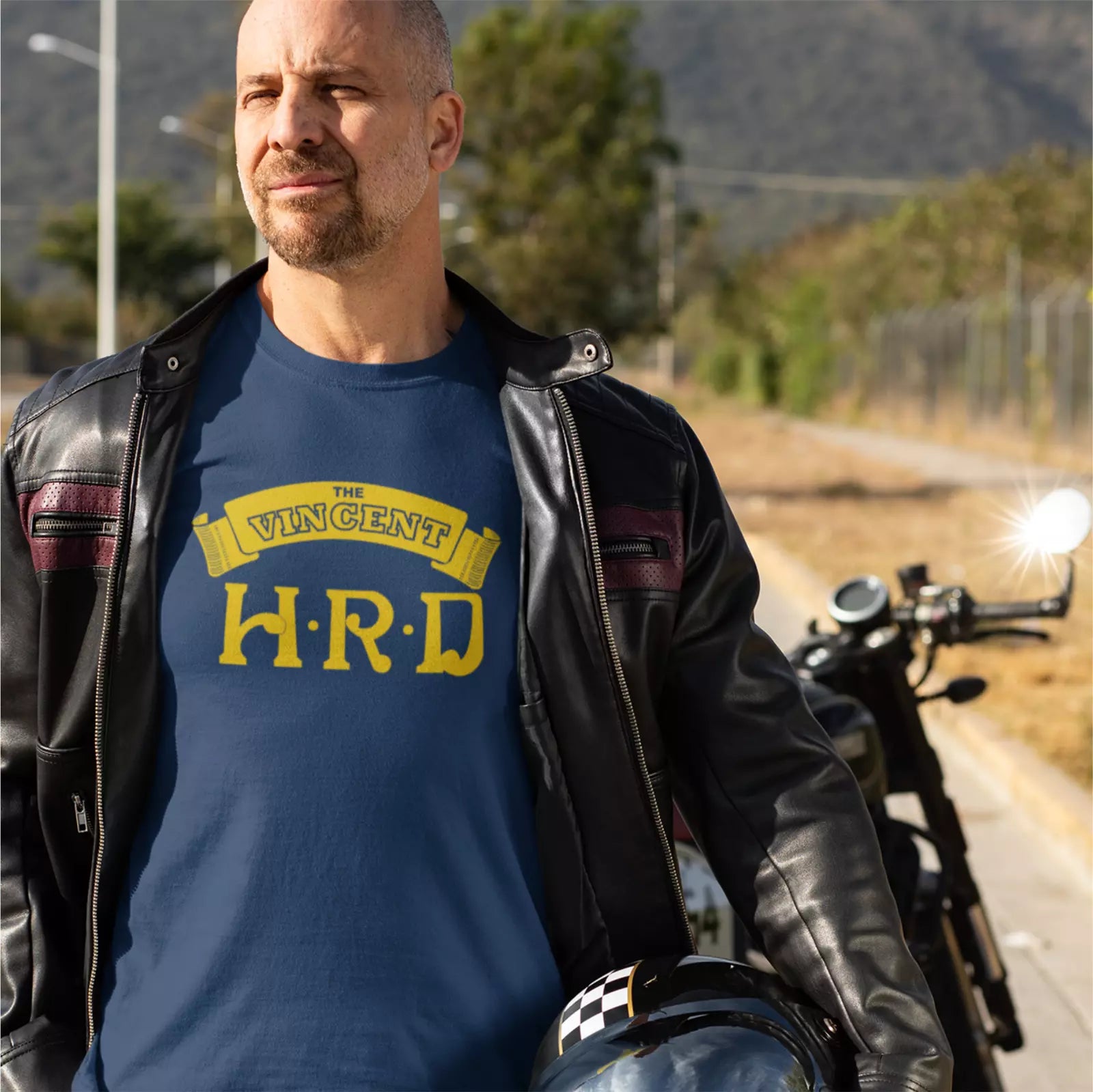 Man wearing a blue t-shirt with 'The Vincent H.R.D' logo, standing next to a motorcycle on a road.