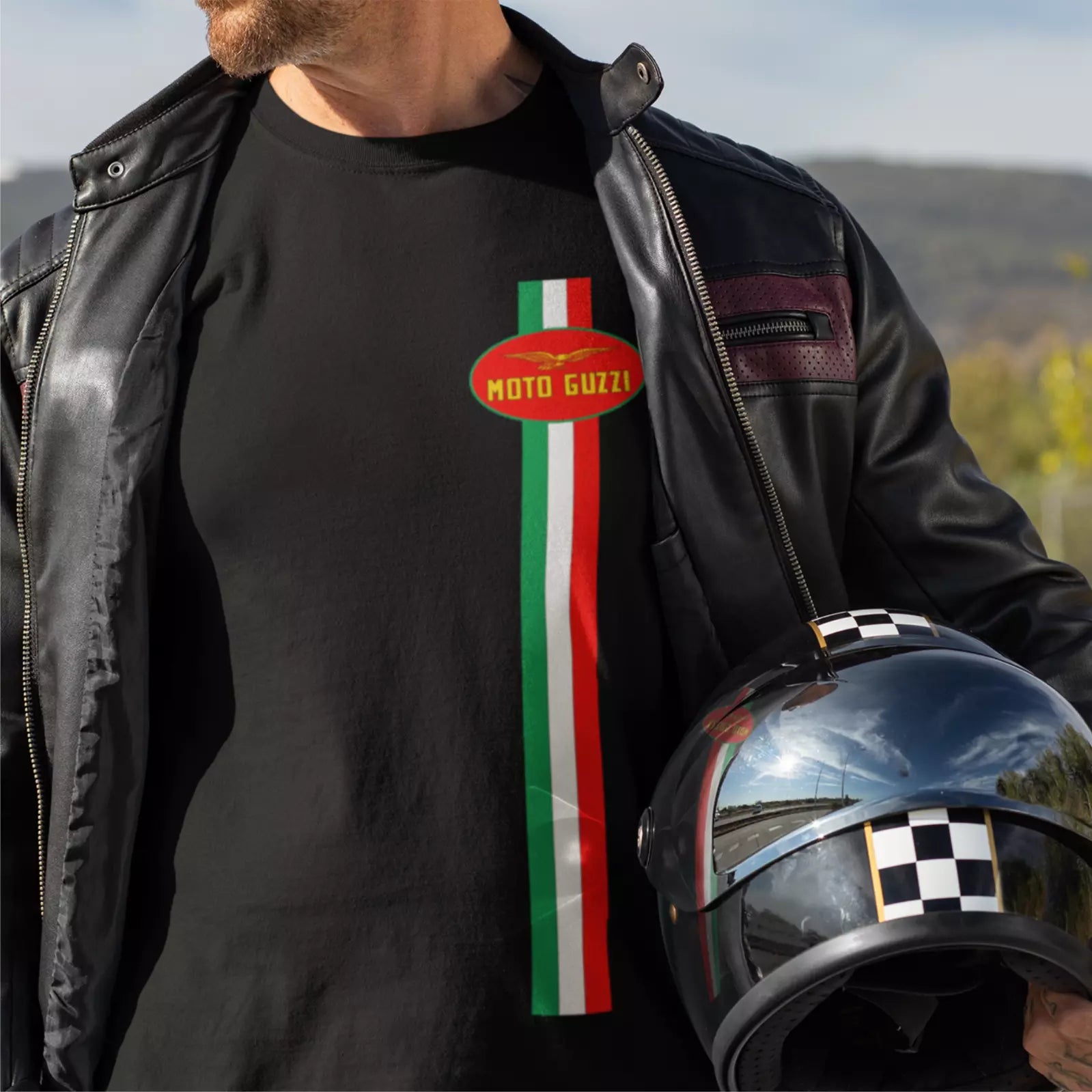 Person wearing a black t-shirt with Moto Guzzi logo and holding a helmet outdoors.