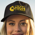 Person wearing a black cap with yellow text 'I LIKE TO GET HIGH AT THE WEEKEND' against a light blue sky.
