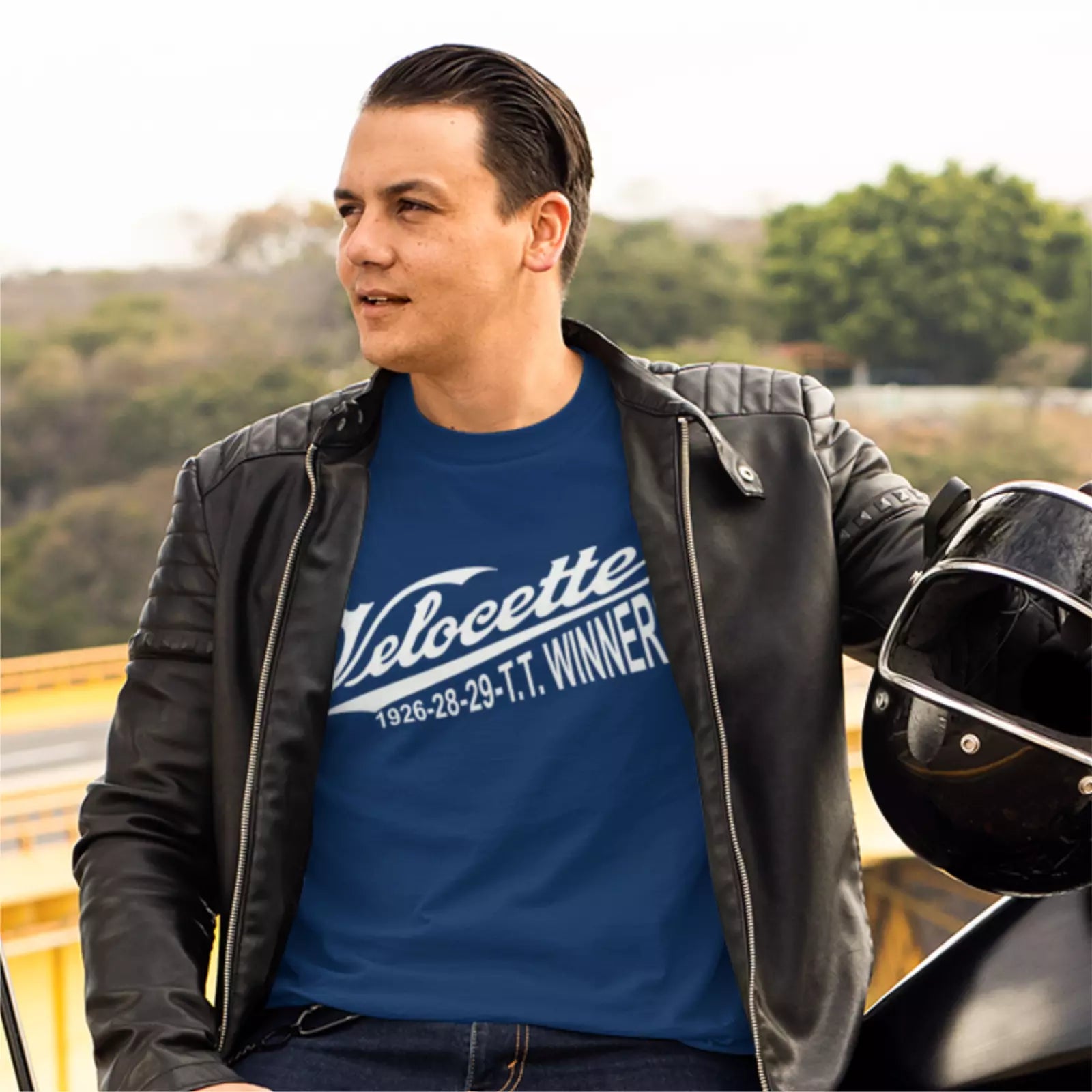 Man wearing a blue t-shirt with 'Velocette' text, sitting next to a motorcycle helmet outdoors.