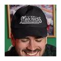 Man wearing a black cap with 'Matchless' logo in front of a green wall with framed pictures.